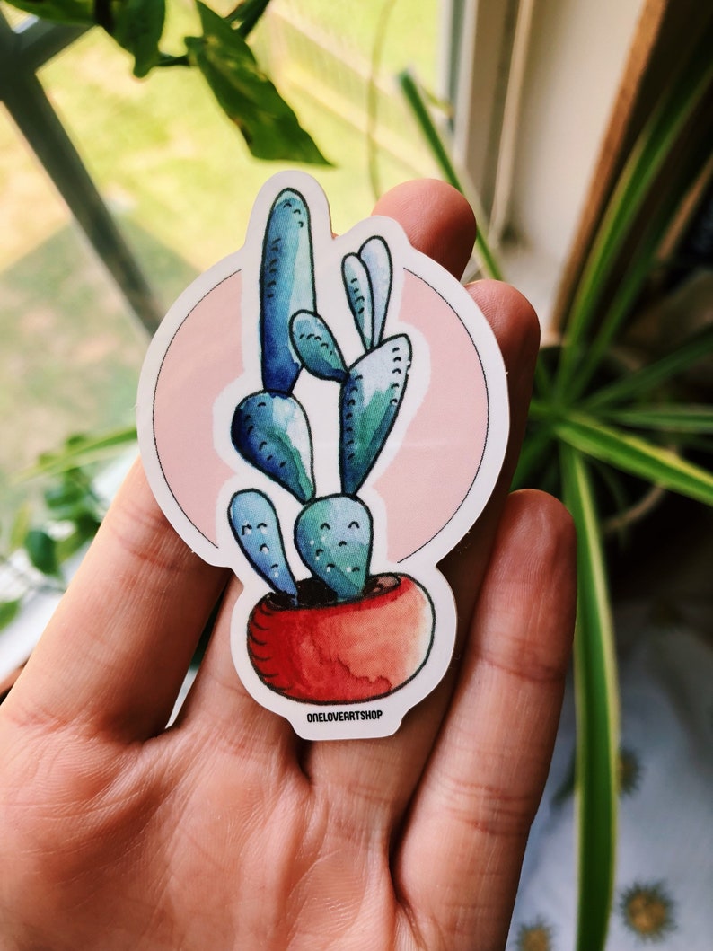 Puede incluir: Una pegatina con una ilustraci&oacute;n de acuarela de un cactus azul en una maceta roja, sobre un fondo rosa. La pegatina tiene un borde blanco y el texto "oneloverartshop" en la parte inferior.