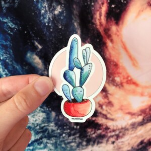 Puede incluir: Una pegatina de un cactus azul en una maceta roja con un c&iacute;rculo rosa alrededor. La pegatina tiene el texto "oneloveartshop" en la parte inferior.