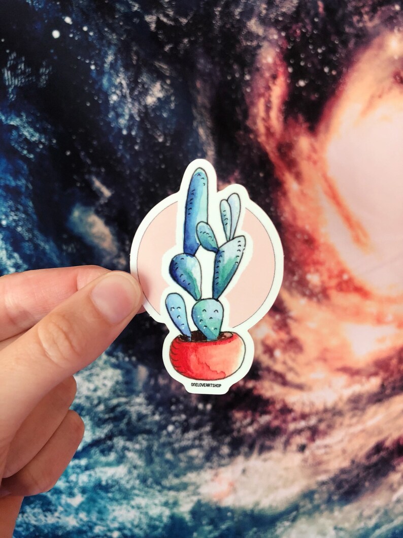 Puede incluir: Una pegatina de un cactus azul en una maceta roja con un c&iacute;rculo rosa alrededor. La pegatina tiene el texto "oneloveartshop" en la parte inferior.