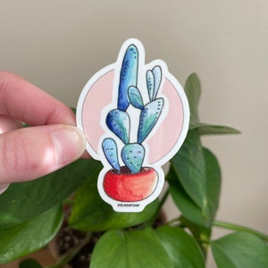 Puede incluir: Una planta de cactus azul y verde en una maceta roja con un fondo rosa. La pegatina tiene el texto "oneloveartshop" en la parte inferior.
