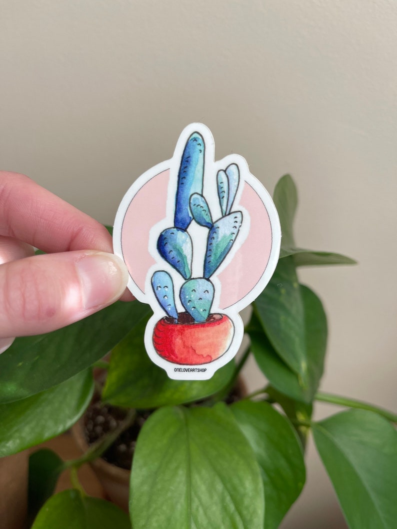 Puede incluir: Una planta de cactus azul y verde en una maceta roja con un fondo rosa. La pegatina tiene el texto "oneloveartshop" en la parte inferior.