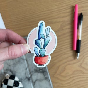 Puede incluir: Una planta de cactus azul y verde en una maceta roja con un c&iacute;rculo rosa detr&aacute;s. La pegatina tiene el texto "oneloveartshop" en la parte inferior.
