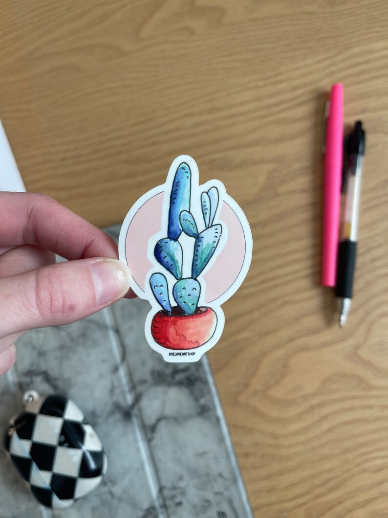 Puede incluir: Una planta de cactus azul y verde en una maceta roja con un c&iacute;rculo rosa detr&aacute;s. La pegatina tiene el texto "oneloveartshop" en la parte inferior.