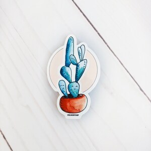 Puede incluir: Una planta de cactus azul en acuarela en una maceta de terracota con fondo rosa. La pegatina tiene el texto "oneloverartshop" en la parte inferior.