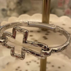 May include: A silver bracelet with a textured cross design. The bracelet is open and has a spring clasp.