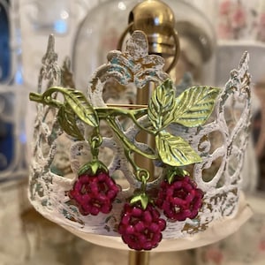 May include: A decorative, ornate crown-shaped object with a distressed white finish. A branch with green leaves and three dangling, beaded, magenta-colored raspberries is attached to the crown. The crown is mounted on a gold-colored stand.