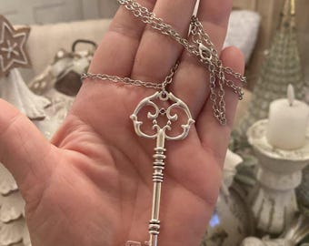 May include: A silver-colored key pendant necklace. The key has an ornate design on the top and a detailed key bit at the bottom. The necklace chain is also silver and is draped over a hand.