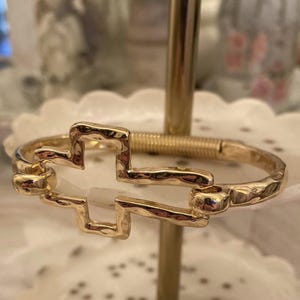 May include: Gold-toned bracelet featuring a cross design. The bracelet has a hammered texture and a hinged closure. The cross is open in the center. The bracelet is displayed on a gold stand.