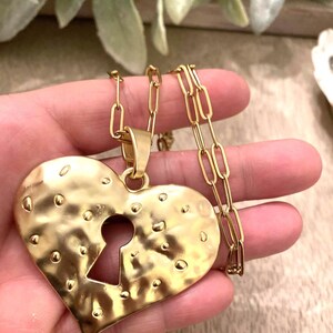 May include: Gold-toned heart pendant necklace with a keyhole design. The heart has a hammered texture and is suspended from a delicate chain. The necklace is a statement piece, perfect for adding a touch of elegance to any outfit.