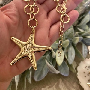 May include: Gold-toned starfish pendant necklace. The necklace features a large, textured starfish pendant suspended from a chain with circular links. The chain includes an adjustable extender. The necklace is a beach-themed accessory.
