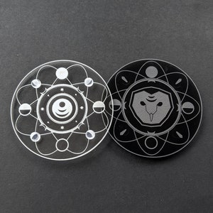 May include: Two round, clear acrylic coasters with a black and white design. One coaster features a stylized owl with a moon and stars. The other coaster features a stylized atom with a moon and stars.