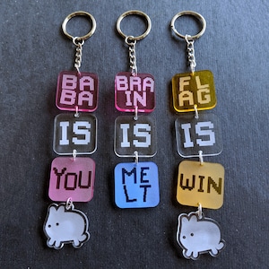 May include: Three keychains with colorful acrylic charms. The top charms read "BA BA", "BRAIN", and "FLAG". The middle charms read "IS", "IS", and "IS". The bottom charms read "YOU", "MELT", and "WIN".