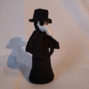 May include: A black crocheted plague doctor doll with a white beak and black button eyes. The doll is wearing a black hat and a long black robe.