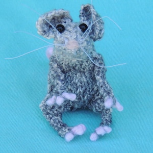 May include: A gray and white knitted mouse with pink paws and black eyes. The mouse is sitting on a blue surface.