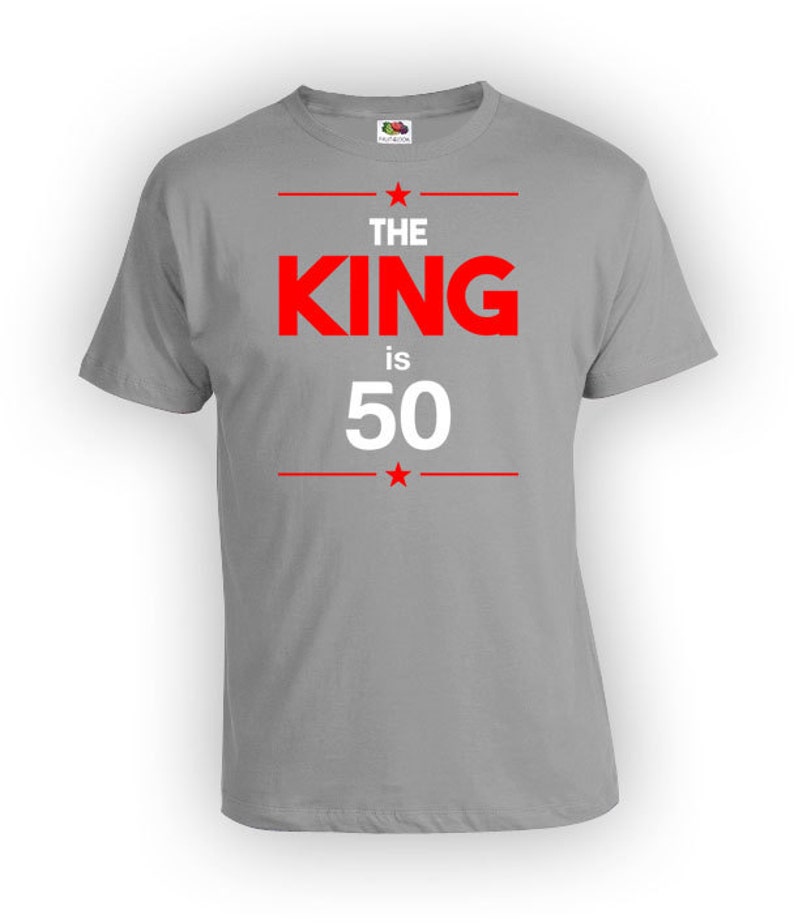 May include: A gray t-shirt with the text "THE KING is 50" in red and white. The text is centered on the front of the shirt, with red lines and stars above and below the text.