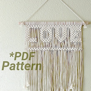 May include: A white macrame wall hanging with the word "LOVE" spelled out in the center. The hanging is attached to a wooden dowel rod and has a fringe at the bottom. The text "*PDF Pattern" is written in green at the bottom left of the image.