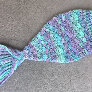 May include: A crocheted mermaid tail blanket in shades of teal, purple, and green. The tail is made of a textured yarn and has a scalloped edge.