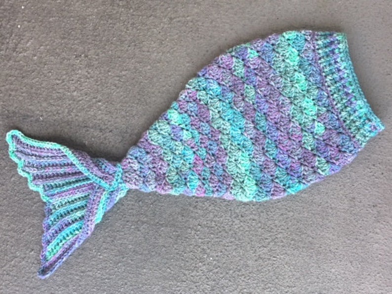 May include: A crocheted mermaid tail blanket in shades of teal, purple, and green. The tail is made of a textured yarn and has a scalloped edge.