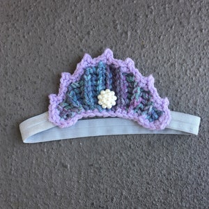 May include: Purple and blue crocheted headband with a white pearl accent. The headband is made of yarn and has a stretchy elastic band.
