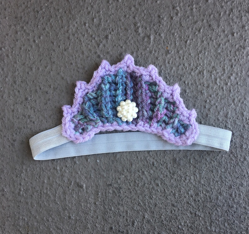 May include: Purple and blue crocheted headband with a white pearl accent. The headband is made of yarn and has a stretchy elastic band.
