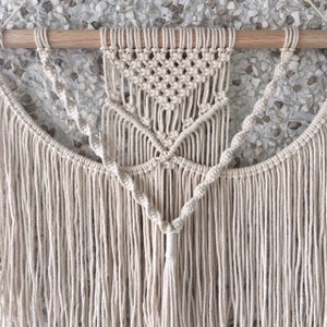 May include: A macrame wall hanging with a geometric design and fringe. The hanging is made of natural cotton cord and is attached to a wooden dowel.