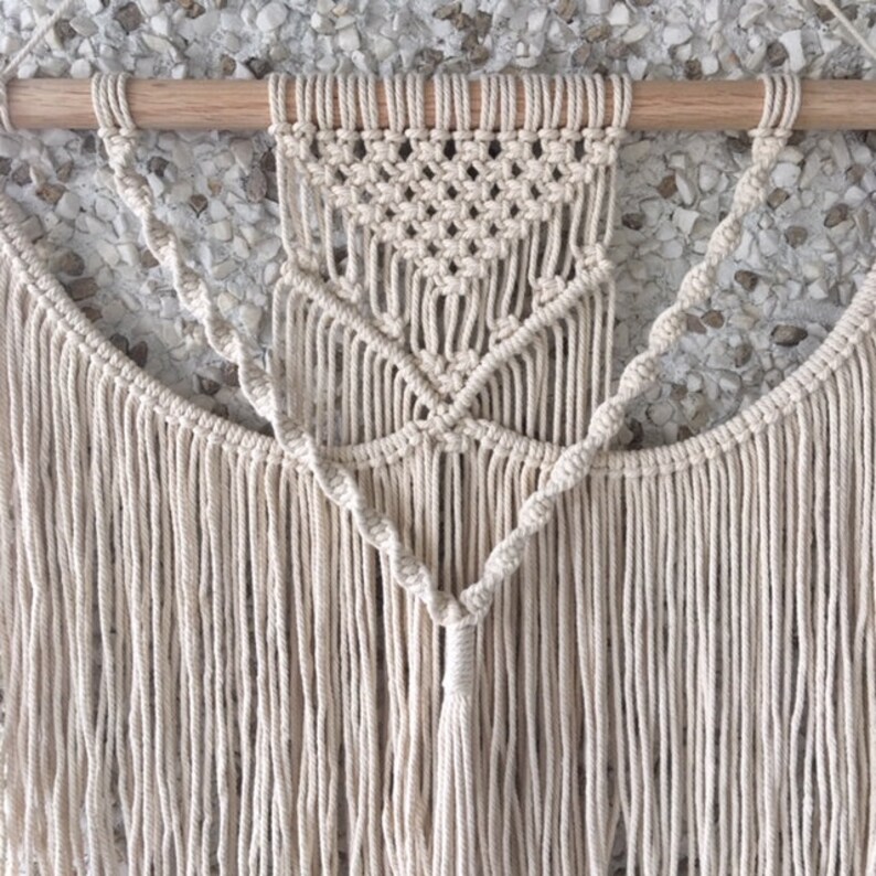May include: A macrame wall hanging with a geometric design and fringe. The hanging is made of natural cotton cord and is attached to a wooden dowel.