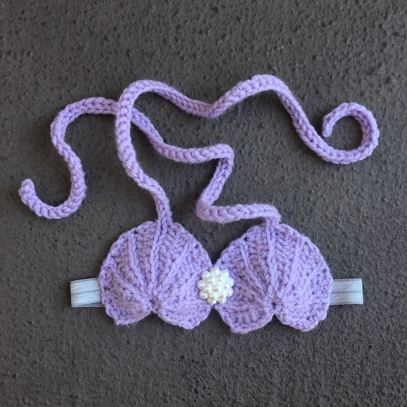 May include: Purple crocheted seashell bra with a white pearl accent. The bra has adjustable straps and a stretchy band.