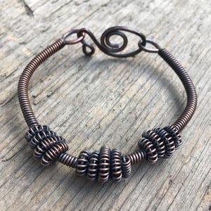 May include: A copper wire bracelet with three coiled wire beads and a spiral design. The bracelet is on a wooden surface.