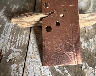May include: A copper-colored metal pendant with a rectangular shape and a curved edge. The pendant has several circular cutouts and a leaf design. It rests on a piece of driftwood against a weathered wooden surface.