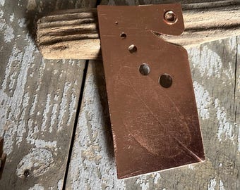 May include: A copper-colored metal piece with various sized holes and a curved edge, resting on a piece of wood. The metal has a slightly textured surface and a small hole near the top. The wood is weathered and has a natural grain.
