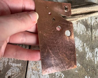 May include: A copper-colored metal piece with a unique shape, featuring several circular holes and a notched edge. The metal has a slightly reflective surface and appears to be hand-crafted. The piece is held by a hand against a weathered wooden background.
