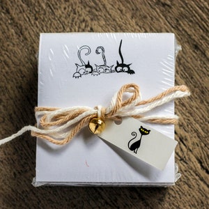 May include: A stack of white note cards with a black and white illustration of cats peeking over the top. The cards are tied with a beige and white twine bow and a gold bell. A small white tag with a black cat illustration is attached to the bow.