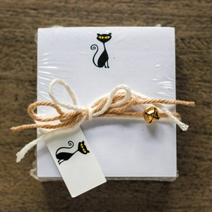 May include: A stack of white notepads with a black cat illustration on the front. The notepads are wrapped with twine and a small gold bell.