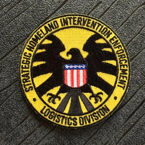 May include: A yellow and black embroidered patch with a black eagle and the text "Strategic Homeland Intervention Enforcement Logistics Division".