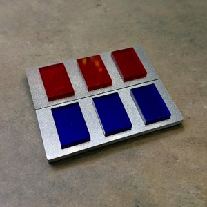 May include: A silver metal badge with six rectangular compartments. The top three compartments are filled with red plastic squares, and the bottom three compartments are filled with blue plastic squares.