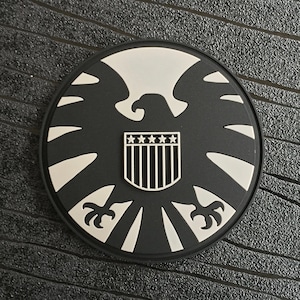 May include: Black and white patch with a stylized eagle design and a shield with five stars and stripes. The patch is circular and has a textured surface.