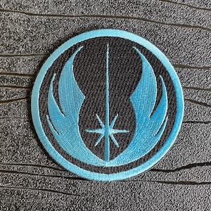Può includere: Una toppa ricamata blu e nera con il simbolo dell'Ordine Jedi. Il simbolo è una spada laser stilizzata con due ali su ogni lato.