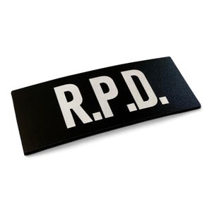 May include: A rectangular black patch featuring the white text R.P.D. in a bold, sans-serif font. The patch has a clean, flat surface with sharp edges and a high-contrast design, displaying the letters clearly against the dark background.
