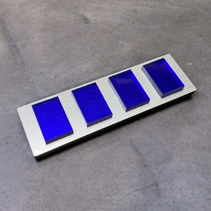 Op de afbeelding: Een zilveren metalen balk met vier blauwe rechthoekige glazen inzetstukken. De balk is een decoratief stuk.