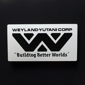 Può includere: Un adesivo rettangolare bianco con la scritta "WEYLAND-YUTANI CORP" sopra un logo "W" nero stilizzato. Sotto, la scritta "Building Better Worlds" tra virgolette. Su sfondo nero.