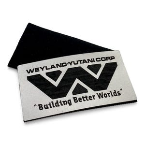 Peut inclure: Une carte rectangulaire en métal argenté avec le texte noir imprimé WEYLAND-YUTANI CORP, un grand logo W noir stylisé et la phrase "Building Better Worlds" placée au-dessus d'une couche de base rectangulaire noire.