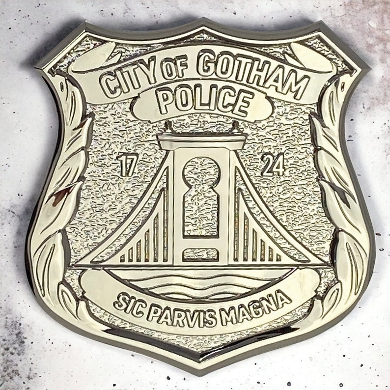 May include: A gold-coloured metal badge with the words "City of Gotham Police" in a curved banner at the top. The badge features a stylised bridge with the numbers "17" and "24" on either side. The words "Sic Parvis Magna" are inscribed in a curved banner at the bottom.
