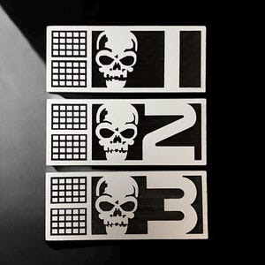 May include: Three silver metal rectangular tags with a black background. Each tag has a grid pattern, a skull graphic, and a number: 1, 2, and 3.