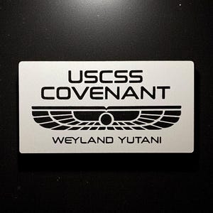 Può includere: Una placca di metallo argentata con testo nero e un simbolo alato stilizzato. Il testo recita "USCSS COVENANT WEYLAND YUTANI".