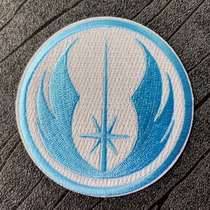 Pode incluir: Um remendo bordado branco com um símbolo azul da Ordem Jedi. O símbolo apresenta duas asas com um sabre de luz e uma estrela no centro.