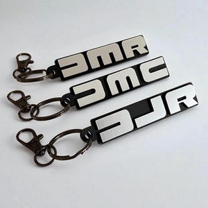 Könnte beinhalten: Drei schwarze Schlüsselanhänger mit silbernen Buchstaben, die "DMR", "DMC" und "DJR" buchstabieren. Jeder Schlüsselanhänger hat einen silbernen Ring und einen bronzefarbenen Verschluss. Die Schlüsselanhänger sind auf einer weißen Oberfläche angeordnet.