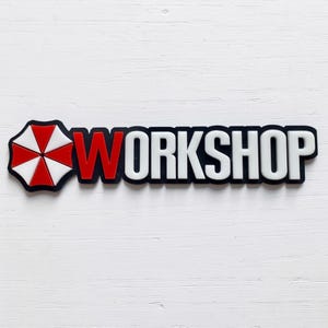 Könnte beinhalten: Ein rechteckiges Schild mit einem rot-weißen, regenschirmförmigen Emblem auf der linken Seite, gefolgt von dem Wort WORKSHOP in fetten, weißen, serifenlosen Großbuchstaben mit einem roten W auf schwarzem Hintergrund.