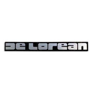 May include: A rectangular emblem featuring the word DE LOREAN in a stylized, bold, silver-toned font. The letters are set against a solid black background, creating a high-contrast appearance for this automotive-style badge or nameplate.