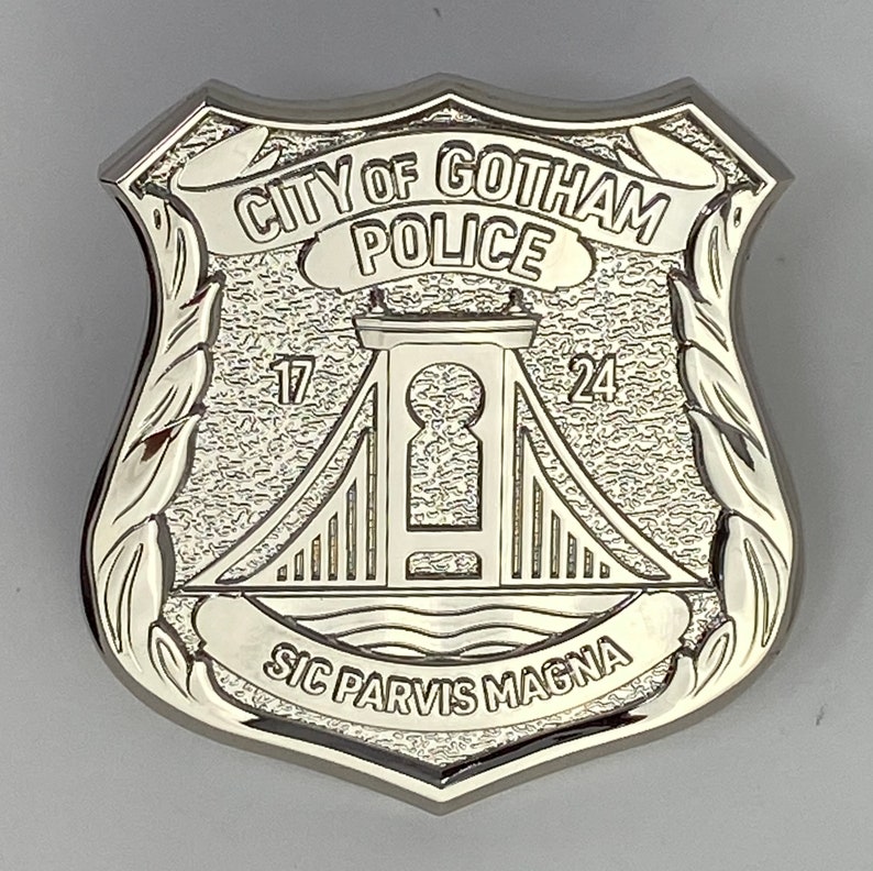 May include: A silver-coloured metal badge with the words "City of Gotham Police" and "Sic Parvis Magna" inscribed on it. The badge features a stylised bridge with a keyhole design.