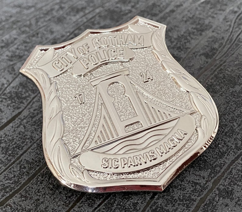 May include: A silver metal police badge with the words "City of Gotham Police" and "Sic Parvis Magna" inscribed on it. The badge features a stylised bridge design with the numbers "17" and "24" on either side.